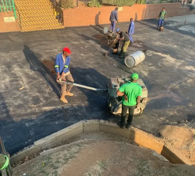 Crew compacting a freshly laid asphalt surface with a walk-behind roller, with the rest of the team working the surface behind them.