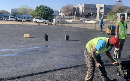 Crew raking hot asphalt at the edge of a newly surfaced parking area, with a roller working behind them.